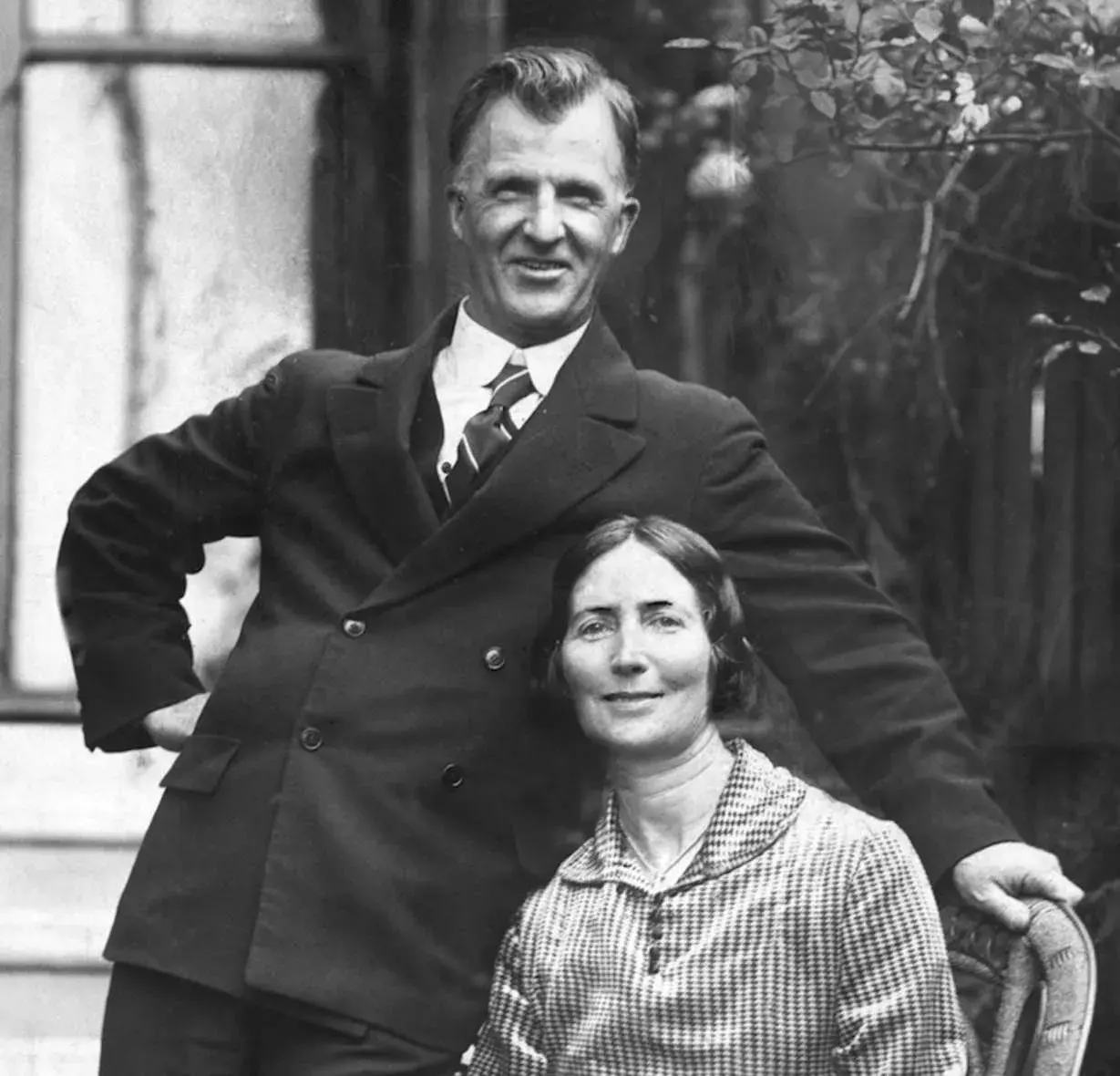 Sarah Scullin sits down while James Scullin stands behind her and leans on her chair and they are both smiling at the camera.  